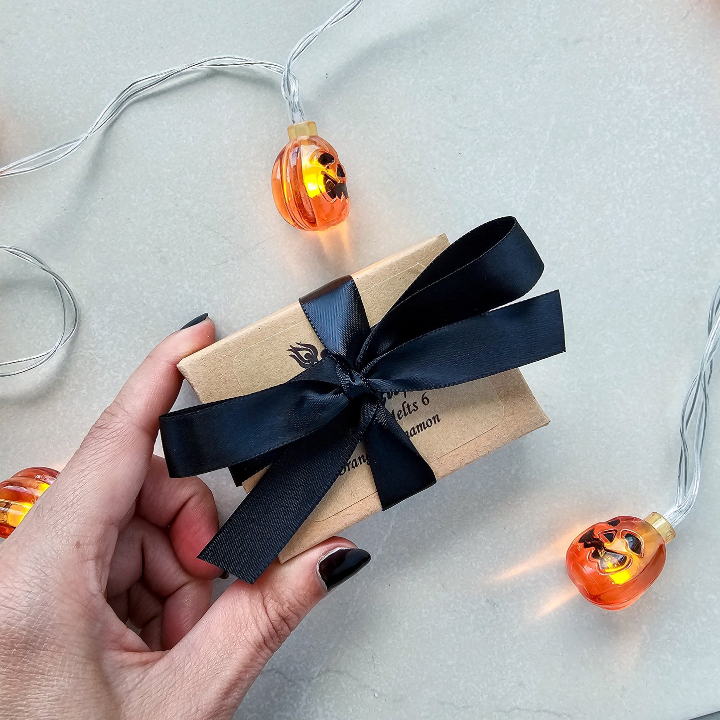 Gift box with black ribbon held by a hand, surrounded by Halloween-themed string lights on a light background