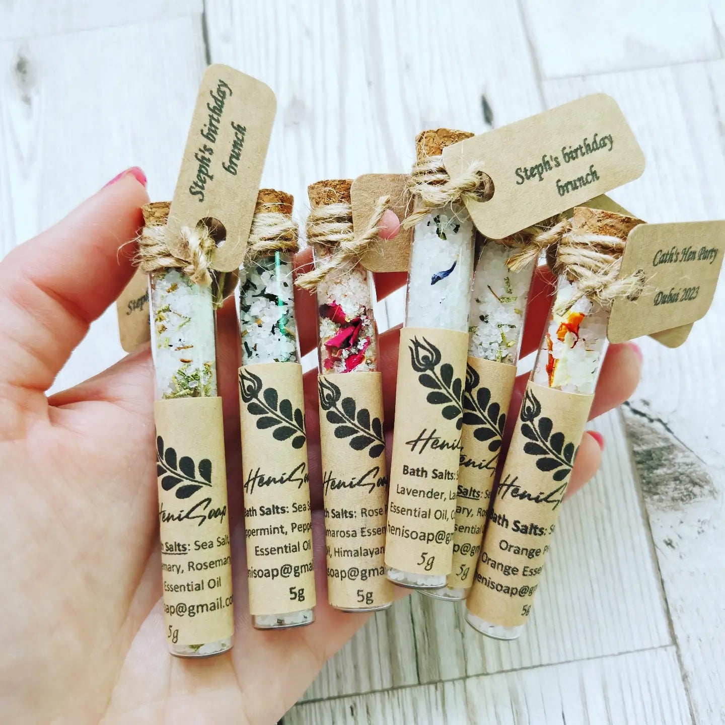 Hand holding small bottles of bath salts with decorative labels on a wooden surface