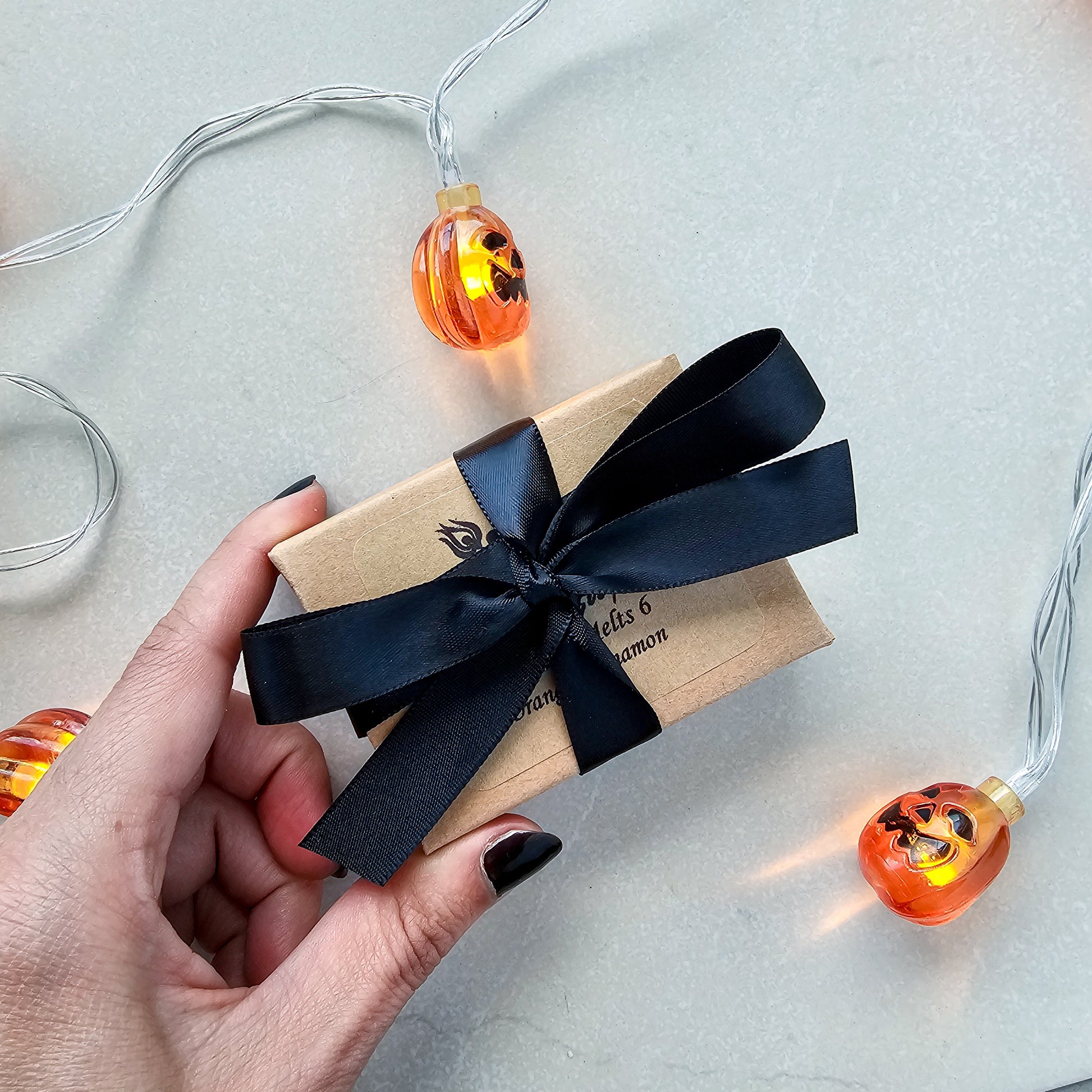 Gift box with black ribbon held by a hand, surrounded by Halloween-themed string lights on a light background