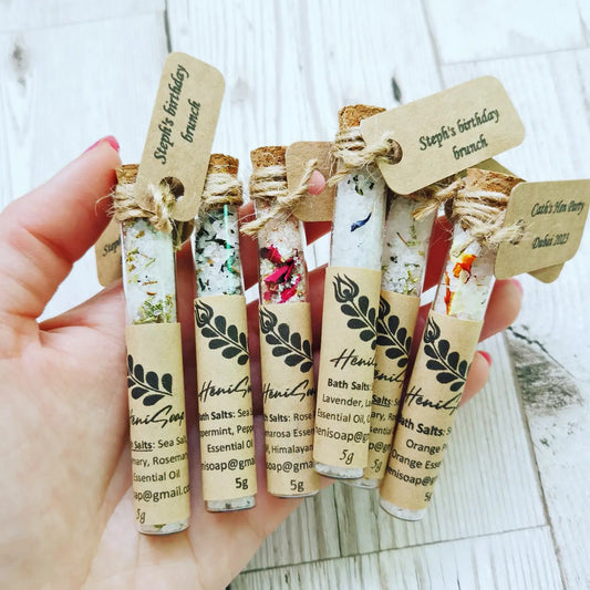 Hand holding small bottles of bath salts with decorative labels on a wooden surface