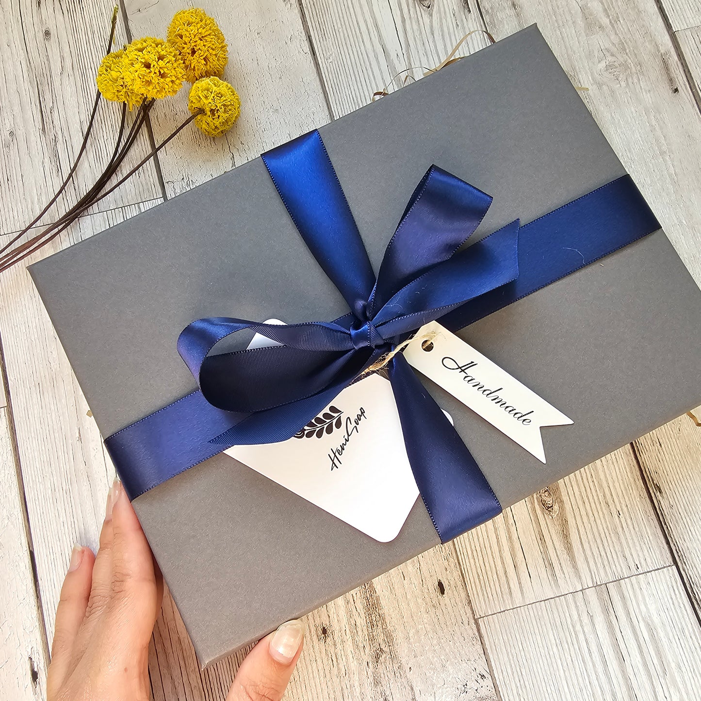 Gray gift box with a blue ribbon held by a hand on a wooden surface.