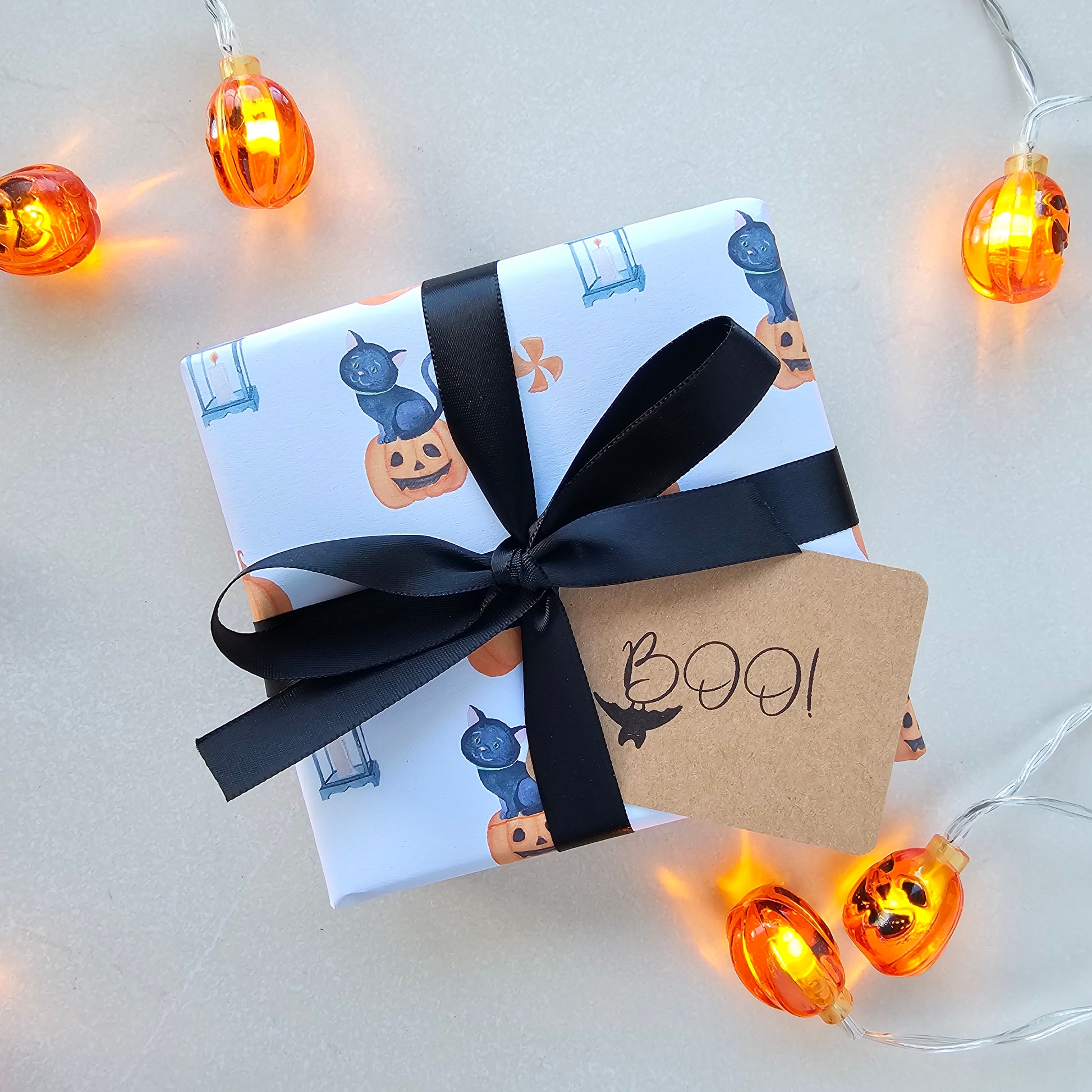 Gift wrapped in Halloween-themed paper with a black ribbon, surrounded by small orange lights on a light background.