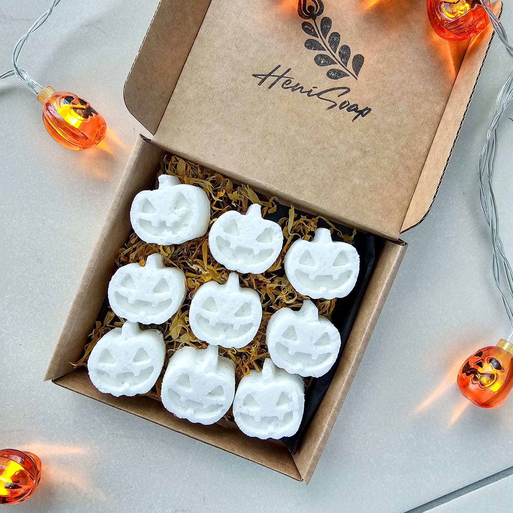 Box of white pumpkin-shaped bath bombs with 'Heni Soap' branding, surrounded by Halloween lights.